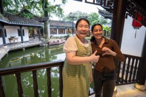 Two women hug and smile on a balcony above a pond