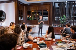 A speaker stands and addresses diners seated at round tables