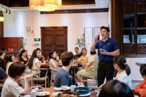 A man with a microphone speaks to a group seated at dinner tables
