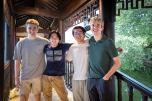 Four friends stand arm in arm on a wooden balcony beside greenery