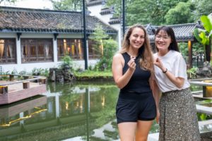 Two women pose beside a pond in front of a traditional building
