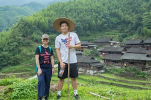 Two people stand on a hillside path above a village and terraced fields