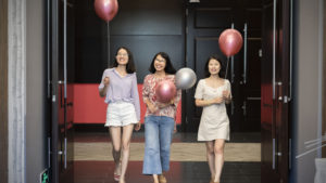 Three women walk through a dark doorway holding balloons