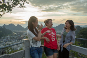 Three friends laugh together at a mountain overlook above the city at sunset