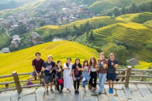 A group poses at a railing above golden rice terraces and a mountain village