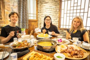 Three friends smile over a hot pot meal with many dishes
