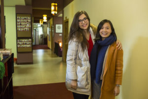 Two women stand together smiling in a warmly lit hallway