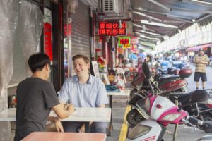 Two friends talk at a sidewalk food stall table