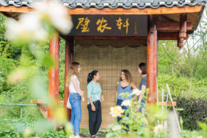 Four women chat under a wooden pavilion behind white flowers