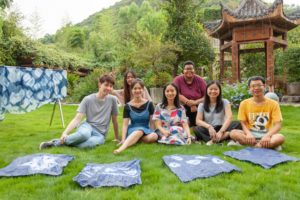 A group sits on the grass behind their blue tie-dye cloths near a pavilion