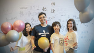 Four people pose with balloons in front of a wall of handwritten messages
