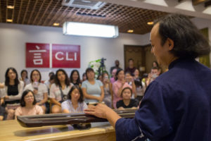 A musician plays a long Chinese zither for a seated audience below a CLI sign