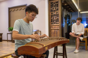 A man in a gray T-shirt plays a guzheng as another man watches
