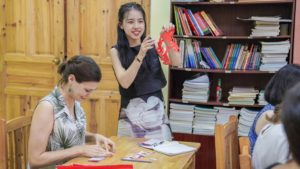 An instructor holds up a finished red paper cut beside a student
