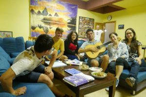 Students relax on blue couches in a yellow lounge, one playing guitar