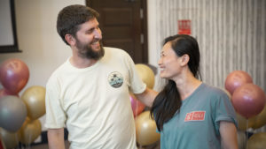 A bearded man and a woman in a CLI shirt laugh together beside party balloons
