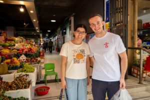 Two people pose together in front of fruit stalls at a market