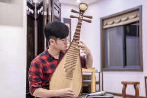 A man in a plaid shirt plays the pipa in a bright room
