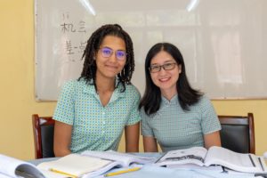 Two women smile at a desk with textbooks in front of a whiteboard