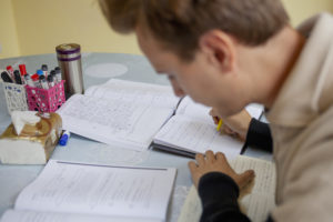 A student writes notes in an open workbook surrounded by study materials