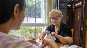 Two women fold sheets of paper together at a table by a window