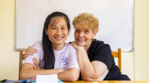A teacher and student smile with arms around each other at a desk