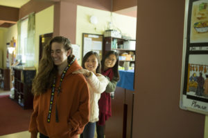 A woman in an orange sweater stands in a hallway with two people behind her