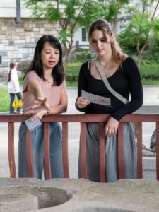 Two women chat at a red railing, one holding a sheet of paper