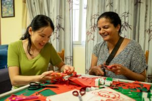 Two women smile while cutting red paper designs at a craft table