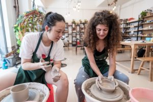 Two women in aprons shape clay on side-by-side pottery wheels