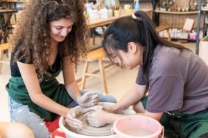 A student shapes clay on a pottery wheel with an instructor's help
