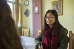 A smiling woman in glasses and a red scarf sits in a classroom