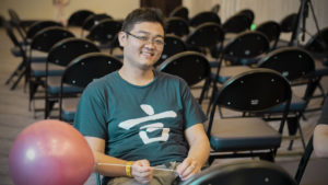 A smiling man sits among rows of folding chairs with a pink balloon