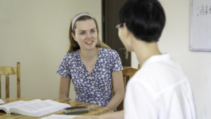 A smiling student listens to her teacher at a desk with an open book
