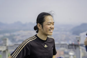 A smiling man in a black sports shirt with a hazy city and hills behind