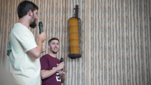 Two men speak into microphones on stage beside a hanging lantern