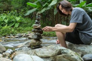 A person crouches beside a creek, carefully stacking stones