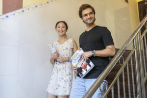 Two people holding books smile beside a staircase