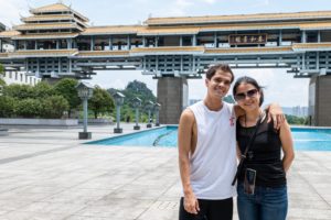 Two people pose by a pool in front of a large covered bridge-style building