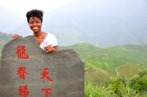 A smiling woman stands behind a carved stone reading Longji Rice Terraces