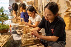 A woman assembles a wooden pagoda model at a crafts table