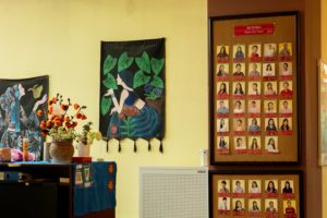 A wall of framed teacher photos beside hanging textiles and flowers
