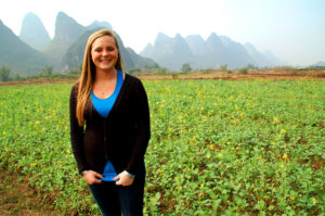A smiling woman stands in a green field with karst peaks behind