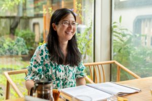 A smiling woman in glasses sits at a table with open books by a window