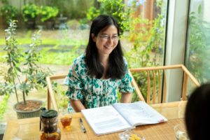 A smiling woman sits with an open textbook at a table by a garden window