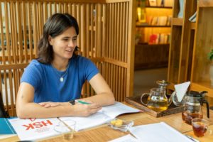A student writes in an HSK workbook beside a tea set