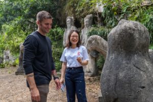 Two people talk among old stone statues in a wooded garden