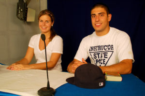 Two people sit behind a microphone at a television studio desk