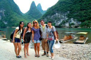 A group stands on a riverbank by bamboo rafts below karst peaks