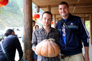 Two men smile with a large pumpkin under a covered walkway